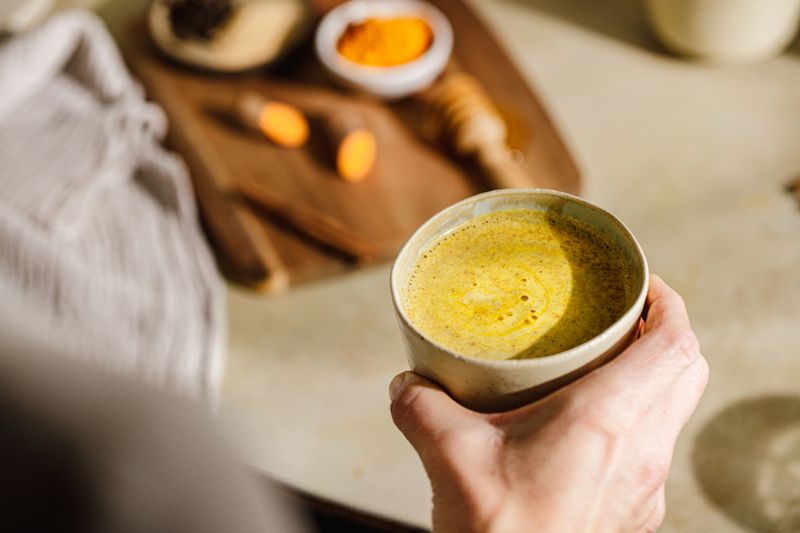 Close-up of a hand holding a ceramic cup filled with golden turmeric latte with ingredients like turmeric root, powder, cinnamon sticks, and honey dipper on a wooden board in the background.
