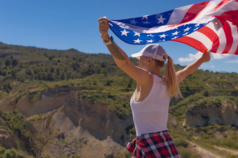 Woman standing outdoors holding an american flag with both hands outstretched, celebrating national pride and independence with a scenic mountain landscape under a clear blue sky