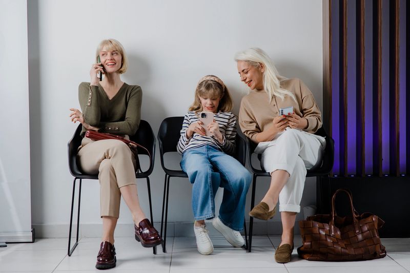 Three females of different ages – a senior woman, an adult, and a young girl – sitting in a waiting room, each focused on her own mobile phone. Scene highlights connecting through technology, showing how digital devices engage all generations in everyday life.
