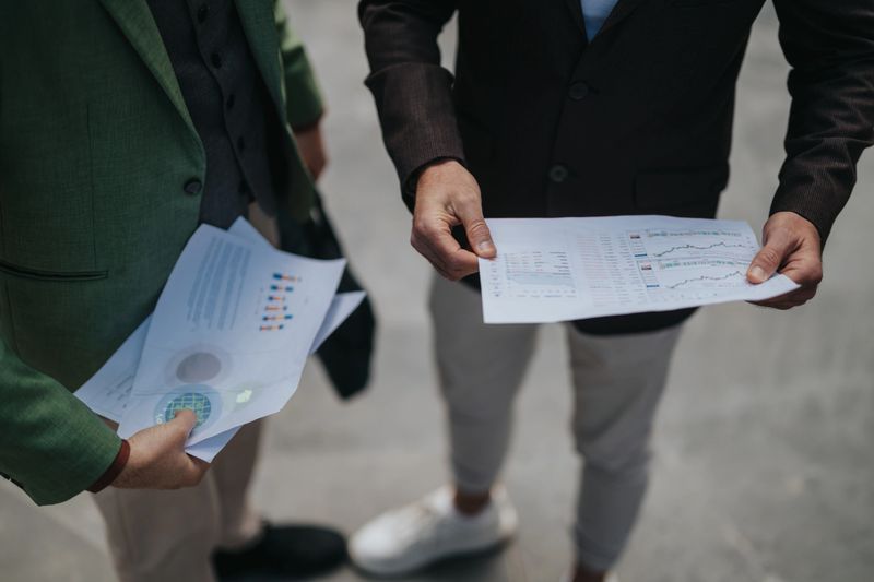 Two colleagues review financial documents and charts during an outdoor business meeting. The partners stand in an urban setting examining printed reports and data.