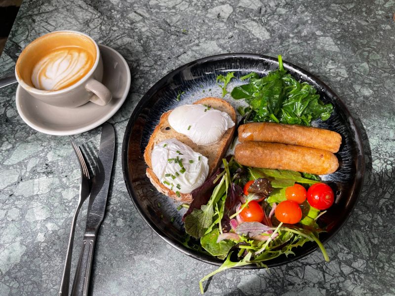 Top-down view of a stylish café breakfast spread on a dark stone plate, featuring poached eggs on toast, fresh green salad leaves, cherry tomatoes, bread rolls, and cutlery, alongside a cappuccino or flat white in a white ceramic cup and saucer on a textured grey concrete surface. Great for food and drink, hospitality, brunch, restaurant, and lifestyle concepts.