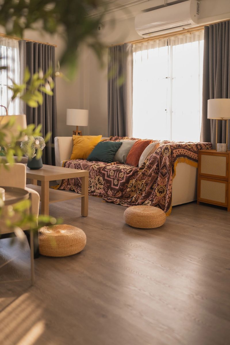 A bright and inviting living room decorated in a modern bohemian style. Featuring a beige sofa with colorful cushions, a patterned throw, and a wooden coffee table. The room is filled with soft natural light from large windows, with green plants in the foreground adding a fresh, organic touch. Concept of home decoration, real estate, and peaceful domestic lifestyle.