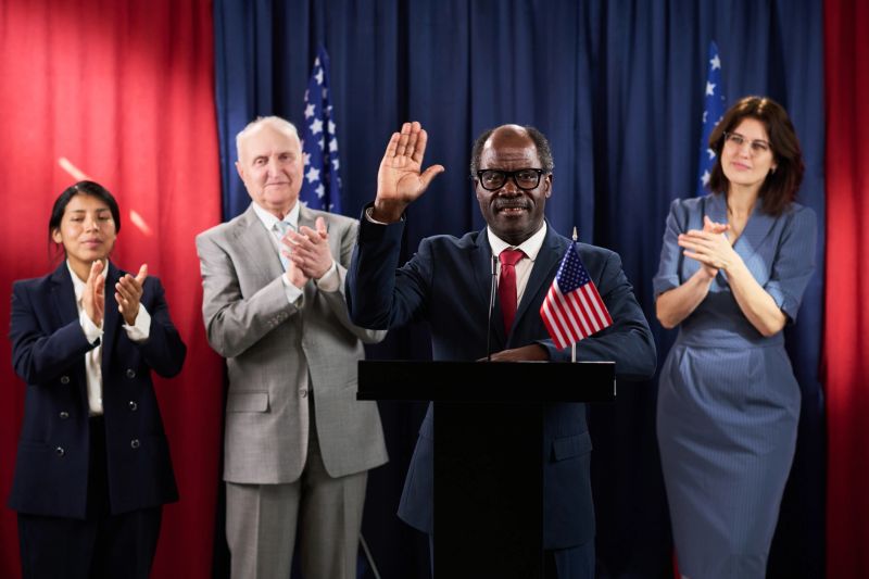 Portrait of African American middle aged politician waving from podium while diverse campaign team clapping during election speech. Ideal for political campaign, leadership