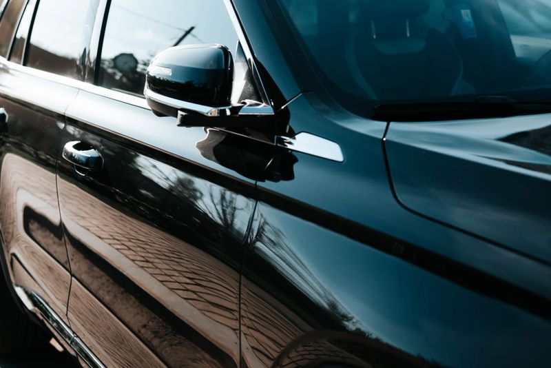 A black car stands parked on a street, reflecting sunlight and nearby surroundings in its polished surface. The scene captures the details of the vehicle and its sleek design under bright light.