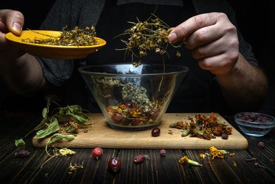 Person preparing dried herbs and flowers in a bowl.