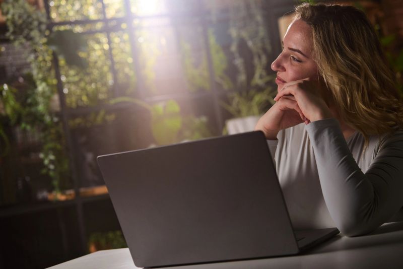 A woman sits at a laptop in a cozy, plant-filled home office, looking thoughtful and relaxed as soft natural light filters through the window behind her, suggesting remote work or creative focus.