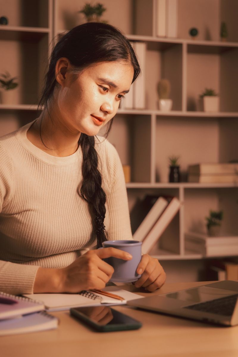 Focused Asian woman working late in a cozy home office, holding a mug while looking at a laptop. Warm lighting highlights productivity, work-life balance, and modern remote professional lifestyle.