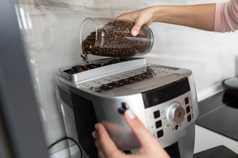 Close up of adult person adding roasted coffee beans into coffee machine. Home kitchen routine, beverage preparation and appliance use concept.