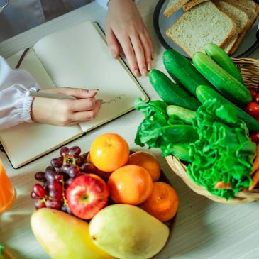 Person writing in a notebook near fresh fruits, vegetables, and bread on a table.