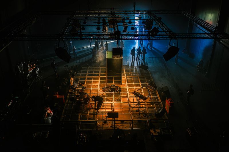 High-angle view of a concert stage with lighting truss, production crew, instruments, and dramatic lighting during live music setup and rehearsal preparation in Bangkok.