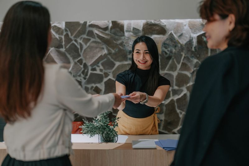 Asian female receptionist hands a brochure to two Asian female guests during consultation or check-in at a luxury reception counter in Kuala Lumpur.