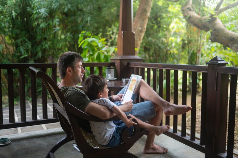 A father and his young son sit comfortably on a wooden rocking chair on a serene porch, surrounded by lush greenery. The father reads a book while the son leans against him, creating a warm and intimate atmosphere. Soft natural light filters through the trees, enhancing the peaceful setting. This image captures the essence of family bonding and relaxation, making it perfect for themes of parenting, leisure, and outdoor living.