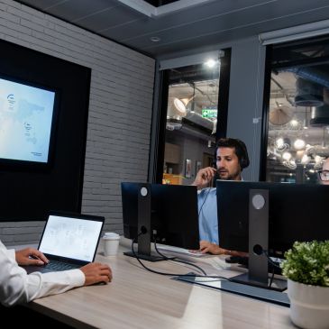 Three colleagues working together in a modern office with computers and a presentation screen.