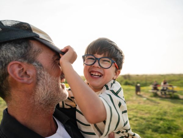 Happy child playfully pulling on an older man's hat outdoors.