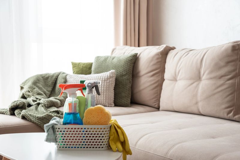 Cozy lifestyle composition featuring a white basket filled with cleaning supplies, including spray bottles, sponge, gloves, and cloth, placed on a white table in a softly lit living room.
