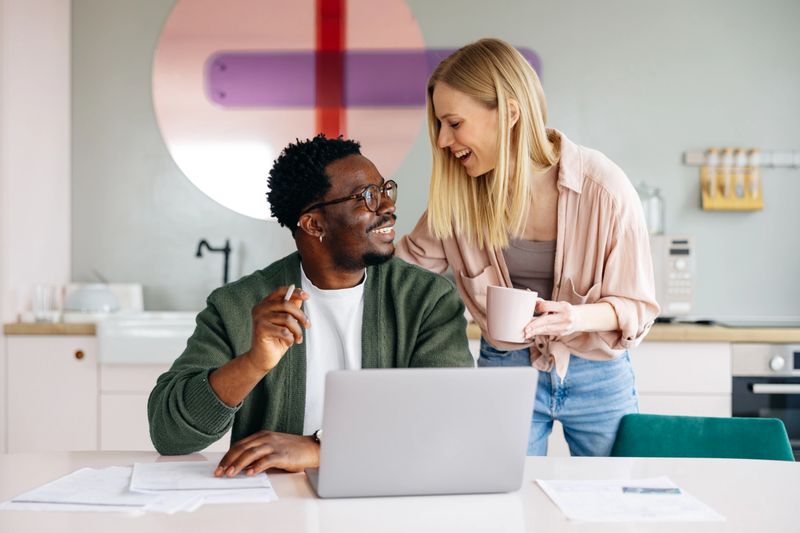 Happy interracial young couple smiling as they manage household finances and taxes together in a cozy modern kitchen with laptop, documents and coffee, planning their budget