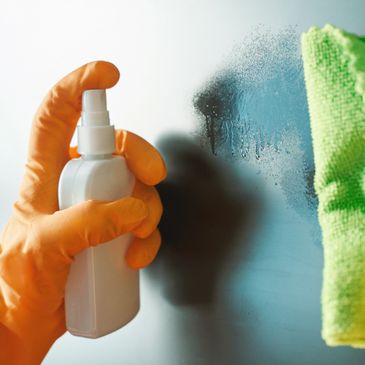 Hand in orange glove spraying cleaner on glass with green cloth.