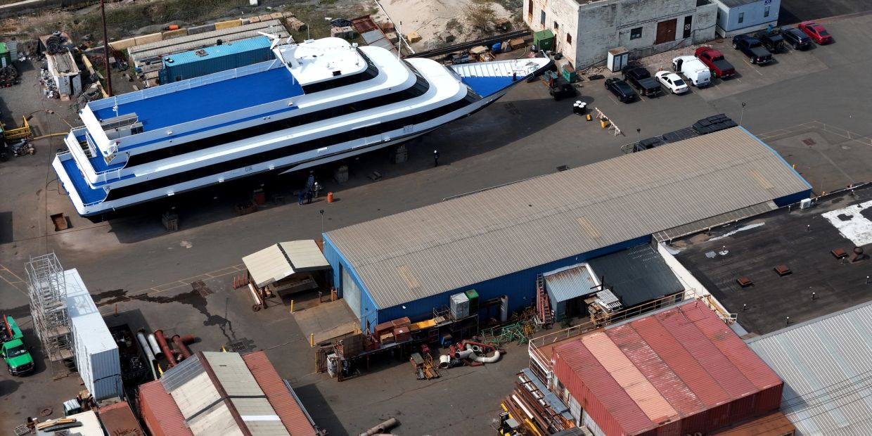 A large white and blue boat docked near industrial buildings and parked cars.