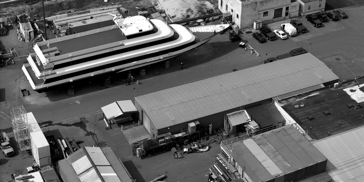 A large white and blue boat docked near industrial buildings and parked cars.