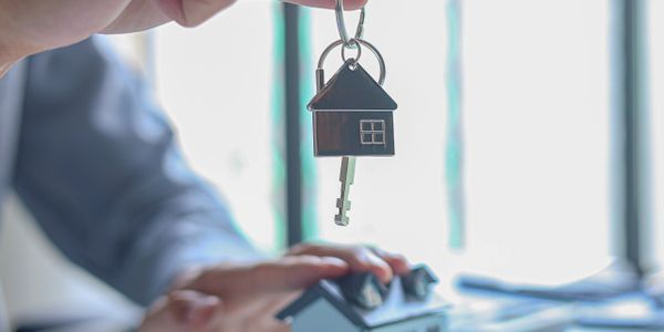 Hand holding a key with a house-shaped keychain over a miniature house model.