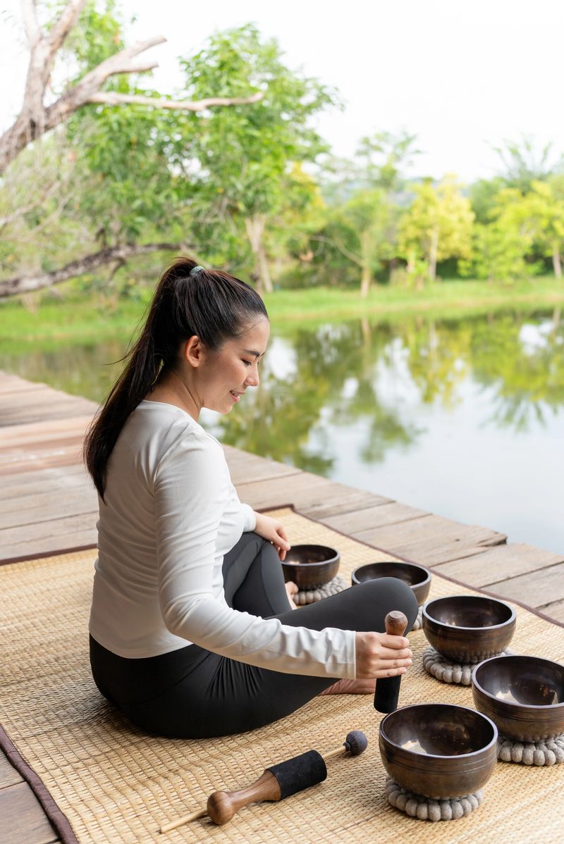 Vertical of asian woman practicing sound bath meditation with Tibetan singing bowl surrounded by peaceful natural scenery at home, inner peace and nervous system regulation for wellness lifestyle.