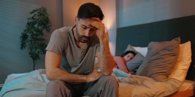 Man sitting on bed looking stressed, woman asleep behind him.