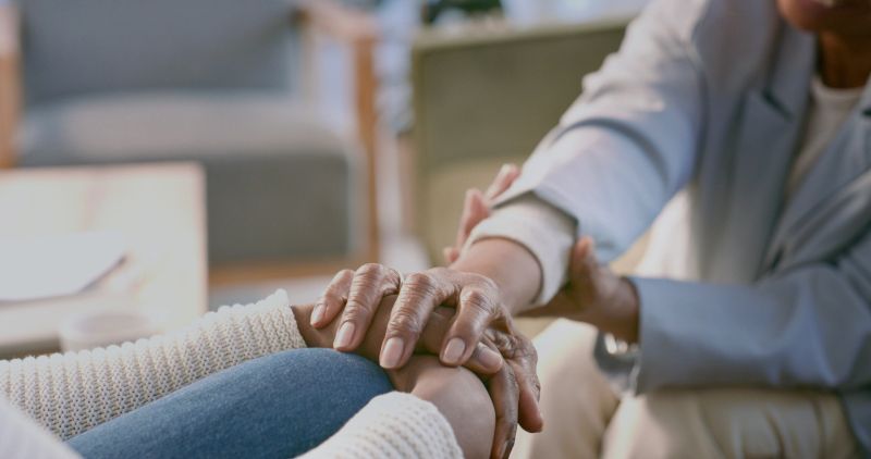 Therapist gently holding a patient's hands in a warm counseling session, conveying comfort, trust, emotional support and compassionate care in a relaxed, home-like clinical setting.