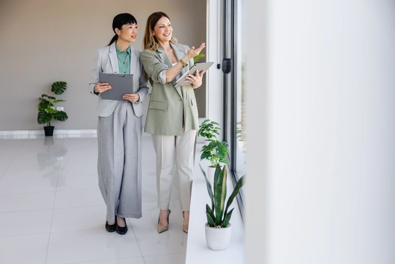 Two professional women stand in a bright, modern office while reviewing documents and a tablet, pointing toward the window during a property tour or client consultation. The scene conveys collaboration, confidence and friendly communication in a clean workplace with natural light and potted plants.