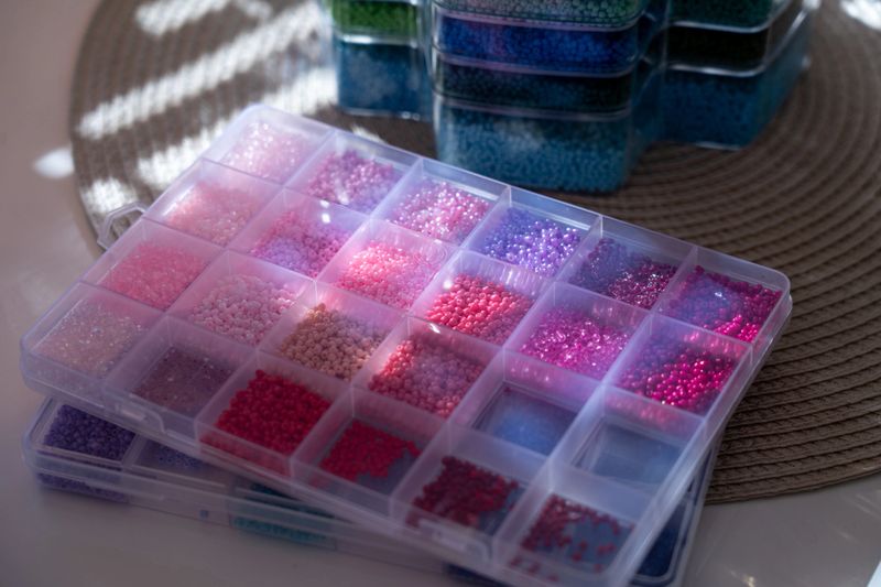 A top-down view of transparent plastic organizers filled with a variety of pink, purple, and blue glass seed beads. The sunlight creates beautiful highlights on the small beads, showcasing a well-organized workspace for jewelry making, embroidery, or handicraft hobbies.