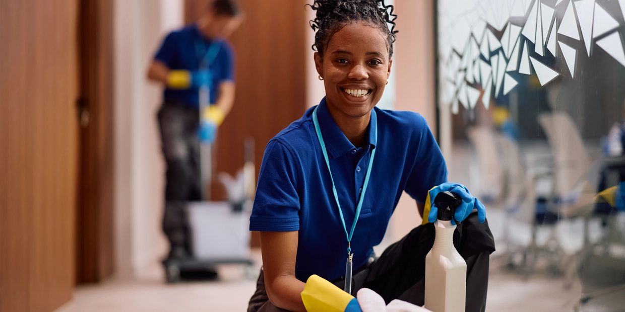 Smiling cleaner in blue uniform holding spray bottle, with colleague cleaning in background.
