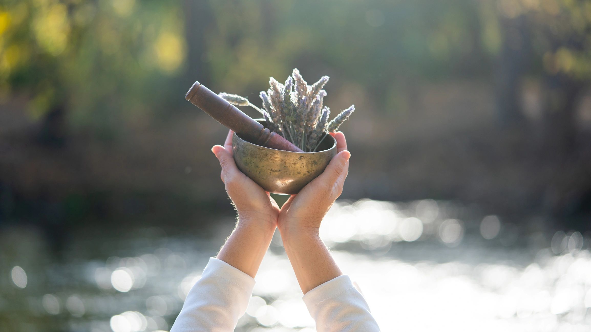 Hands holding a brass bowl with lavender and a wooden pestle outdoors.
