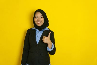 Woman in hijab giving thumbs up against yellow background.