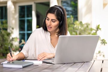 Woman working on laptop outdoors, taking notes with headphones on.