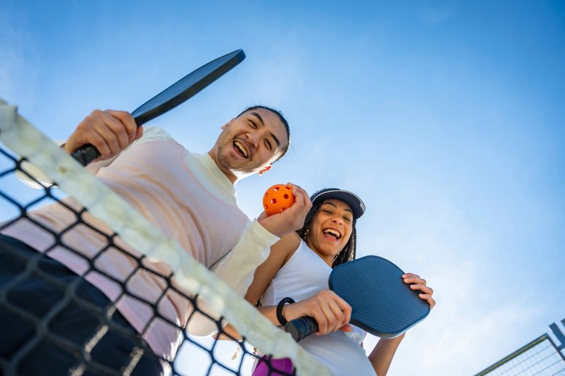 Enthusiastic multi-ethnic players standing on a pickleball court with rackets and a ball, actively enjoying the game and smiling at the camera on a sunny day