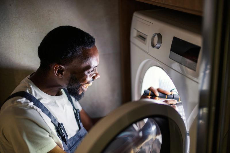 African american repairman inspecting a washing machine with a flashlight, smiling as he performs maintenance and repairs in a home laundry room, professional and focused