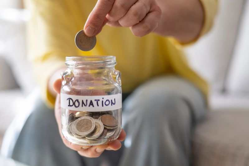 Person holding a glass jar labeled donation filled with coins, dropping another coin into the jar, symbolizing concepts of philanthropy, savings, and financial support