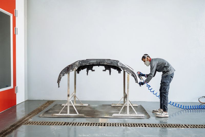 A young man in protective gear is sanding a car body part in a clean workshop environment, showcasing focus and precision in his work.