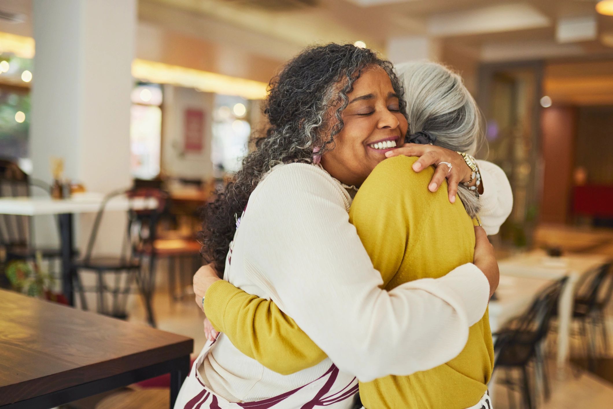 An African American woman with long dark curly hair closes her eyes with a joyful smile while embracing a silver-haired Caucasian friend in a mustard cardigan at an indoor restaurant, capturing a genuine and warm moment of female friendship and connection.
