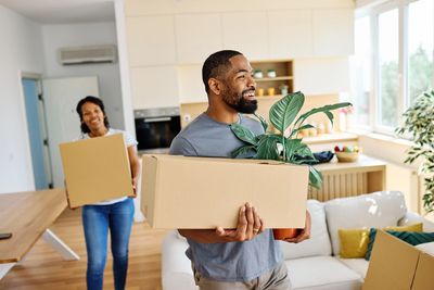 Couple carrying boxes and a plant in a new home.
