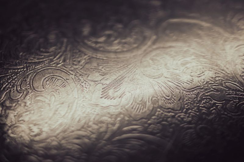 Metallic surface showcasing an ornate floral engraving, featuring a textured silver background with a dark vignette and subtle light reflection, providing a rich, decorative backdrop