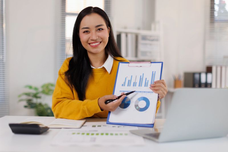 Young adult asian business woman presenting financial data charts and graphs, pointing to statistics on a clipboard while participating in a virtual meeting from her desk in a bright, modern office