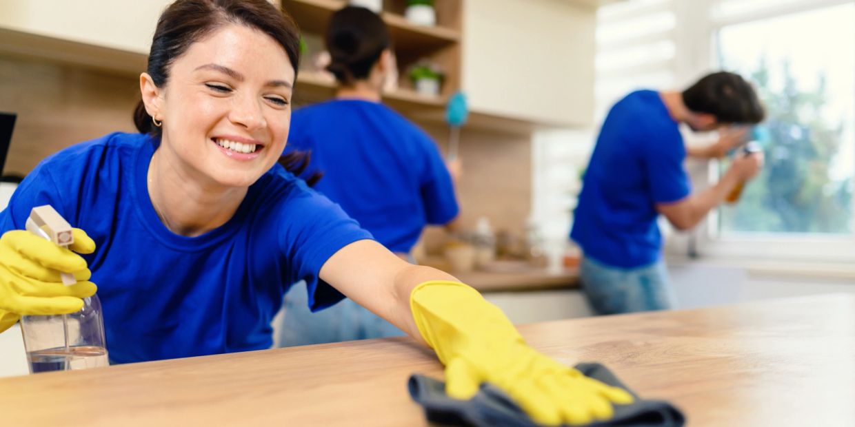 A smiling woman in yellow gloves cleans a wooden surface with others cleaning in the background.