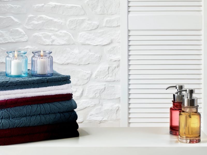 Neatly stacked bath towels in various colors sit on a clean white shelf alongside lit candles and glass soap dispensers. The white textured brick wall in the background adds depth to this calm and elegant bathroom setting. Ideal for home decor, wellness, and interior lifestyle concepts. Horizontal composition.