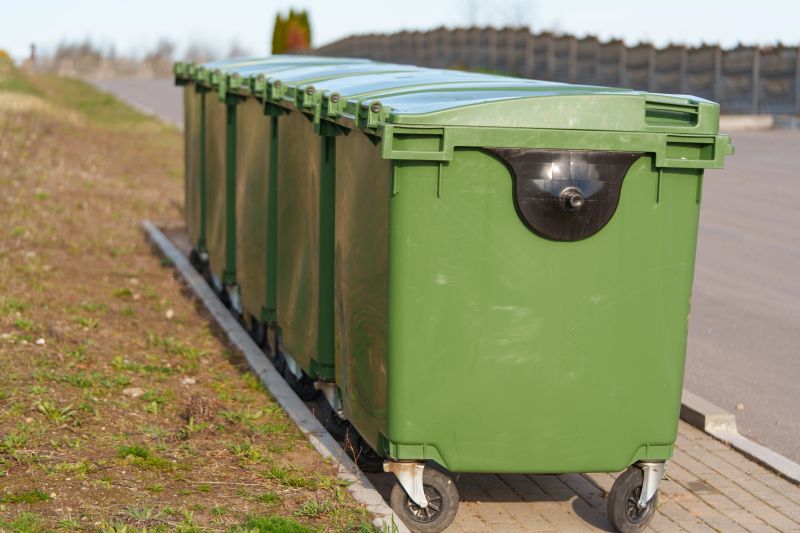 Green plastic garbage containers with 1100-liter capacity, equipped with four swivel wheels, arranged in a row outdoors. The container area is separated from the lawn or walkway.