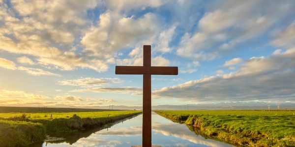 Wooden cross by a calm canal under a dramatic sky.