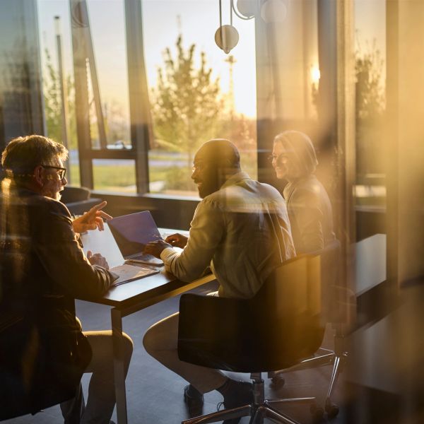 Three people in a meeting with laptops during sunset.