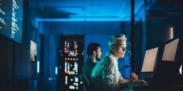Two people working on computers in a dimly lit room with digital clock display.