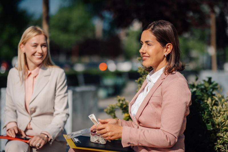 Two smiling adult businesswomen sitting outside, having a refreshing and healthy salad for lunch during their office break