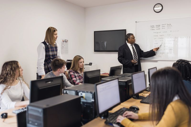 Group of students in a computer class with a teacher explaining on a whiteboard