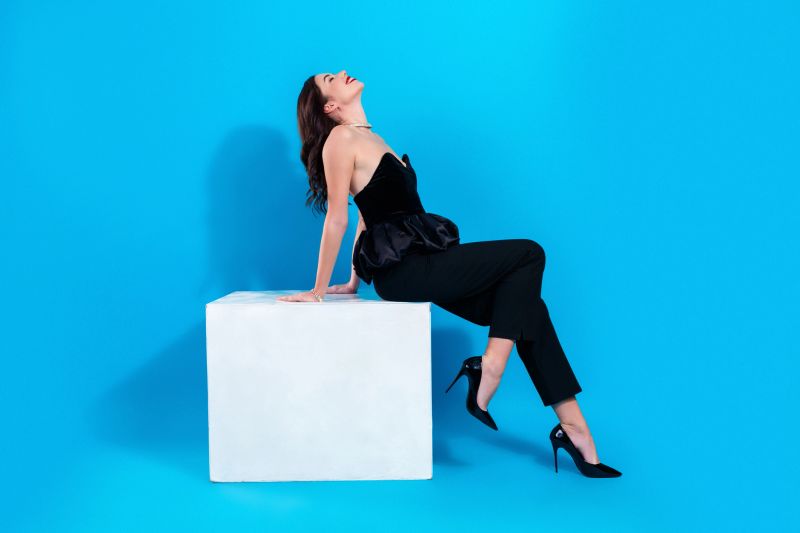 Glamorous fashion model poses confidently on a white block against a vivid blue studio backdrop, wearing a black evening top and trousers with high heels and statement jewelry, conveying elegance and poise.